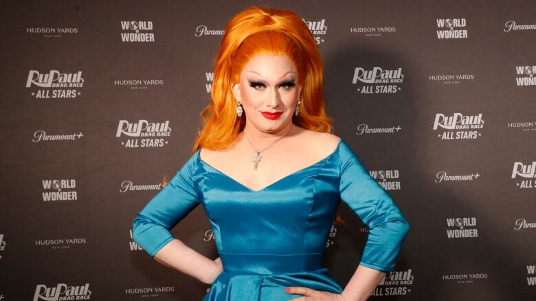 Performer with bright orange hair and a turquoise dress stands on the red carpet.