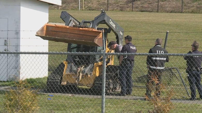 Four police investigators look over a piece of construction equipment, a skid-steer loader, behind a chain-link fence.