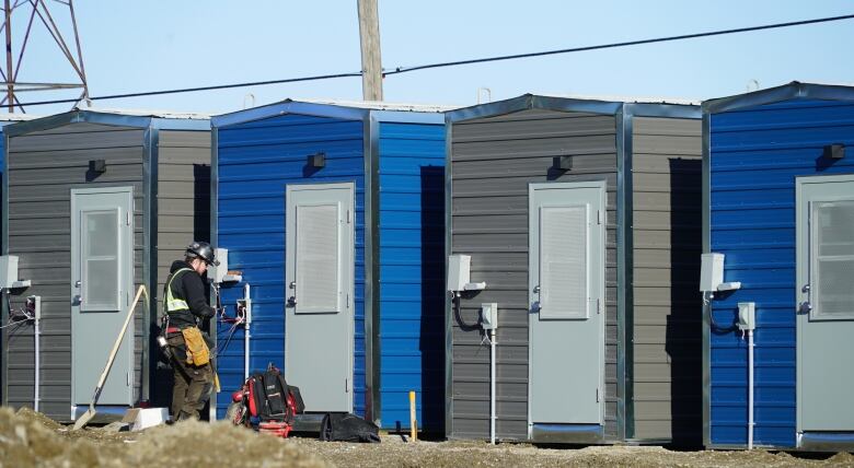 A worker stands in front of tiny homes that are side-by-side. The tiny homes are grey or blue in colour.