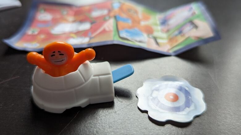 Picture depicts a figurine in an orange suit placed inside an igloo. Next to it is a curved circle with what looks like a curling target inside. Behind the toy is a piece of paper, featuring instructions on how to play it. 