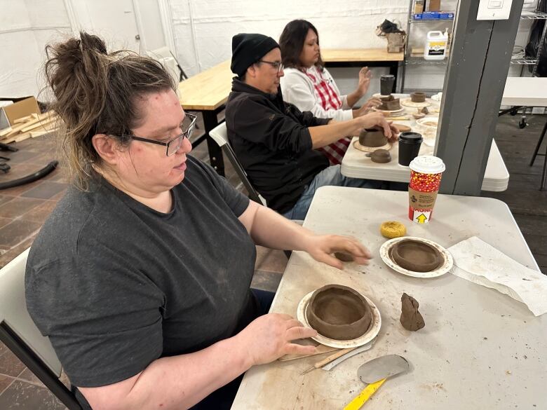 A woman sits at a table, making a small pot out of clay.