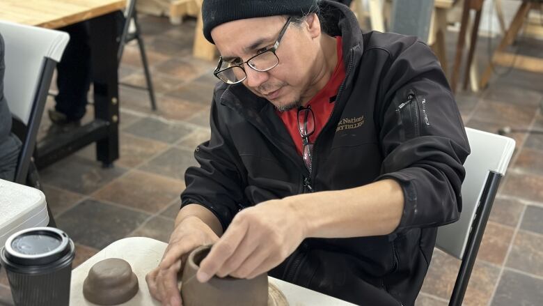 A man sits at a table making a pot from clay.