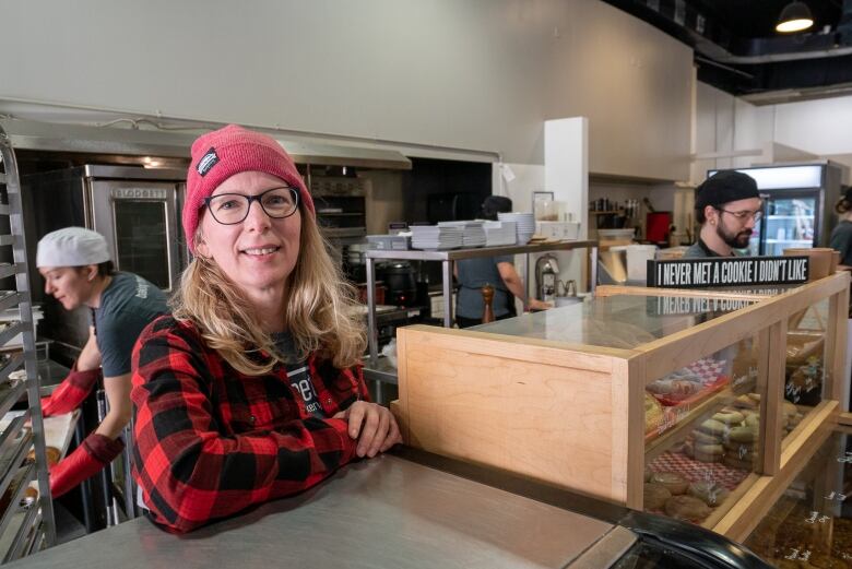 A woman leans on a counter while bakers work behind her.