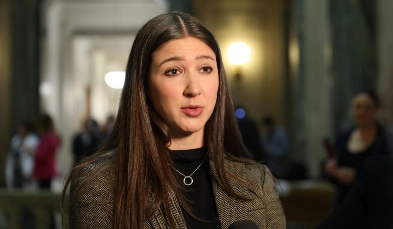 A woman with brown hair, a black sweater and a herringbone jacket speaks with media at the Saskatchewan legislature.