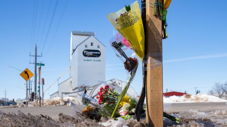 Flowers are strapped to a roadside post, with more flowers at the base of the post, with a white grain elevator in the backdrop that has Gilbert Plains on it.