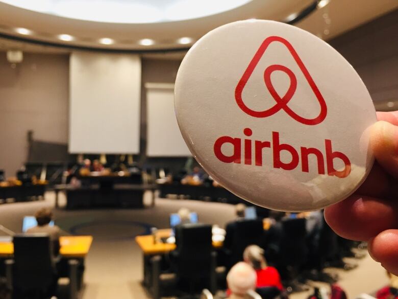 An Airbnb pin is held up against the backdrop of a city council chamber.