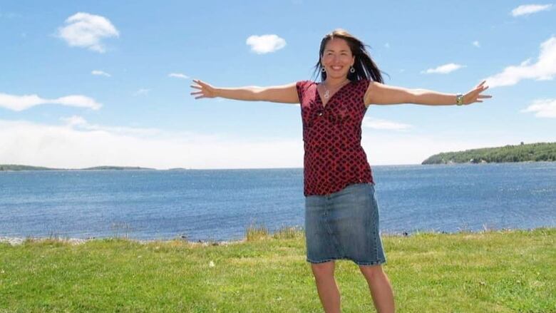 A woman stands on grass with the ocean behind her. She has long dark brown hair and is wearing a red and black top with a denim skirt. Her arms are outstretched and she is smiling.