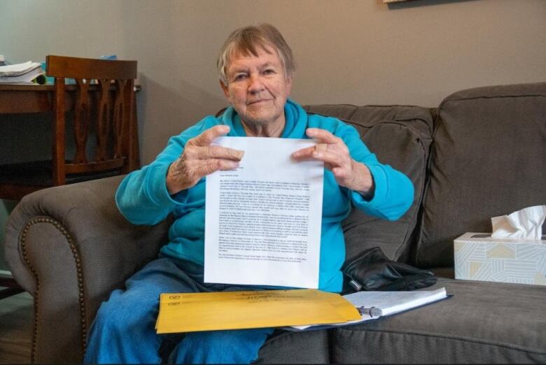 A woman sitting on the couch holds up a piece of paper and looks into the camera.