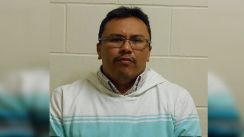 Man in glasses stands straight-faced for photo in white and blue sweater