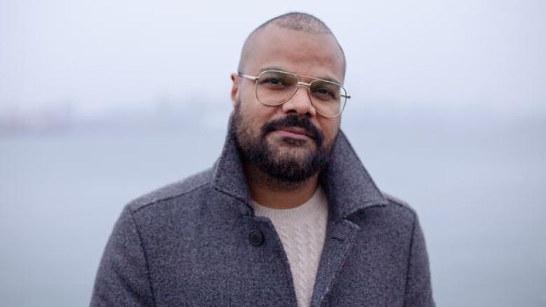 A bearded man with very short hair outside in a beige knitted sweater and grey jacket