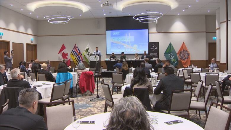 A conference room is pictured with people sitting at tables scattered throughout and a panel of speakers at the front of the room. 