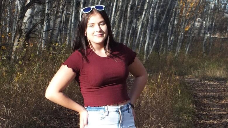 A teenage girl wearing light-coloured blue jeans and a red shirt with sunglasses on her head is pictured standing near a line of trees in the fall. 