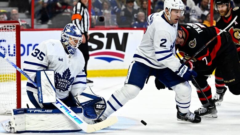 A male ice hockey goaltender stops the puck while down on both knees as two players push against each other in front of him.