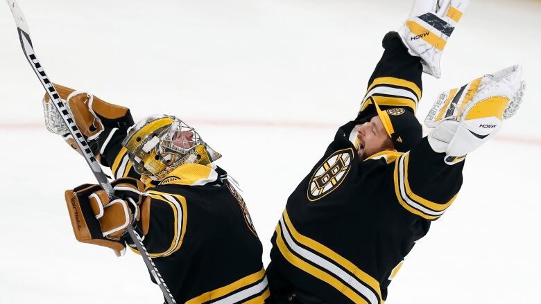 Two goaltenders in black and gold hold their arms skyward pre-embrace. One is wearing a full goaltending helmet and mask and the other wears a baseball cap with a B inside a spoked circle.