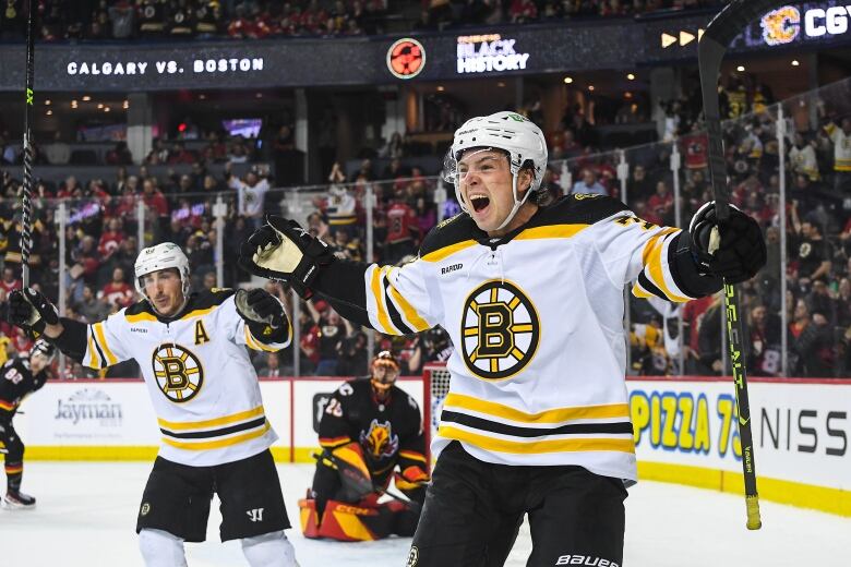 Two hockey players in white jerseys with black and gold spoked Bs on them raise their arms in celebration. In the background a goaltender in a dark uniform is on his knees. Part of the crowd is visible beyond the rink.