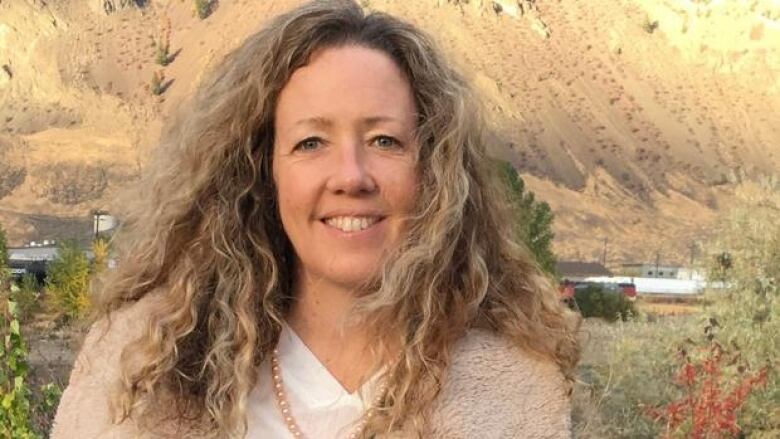 A woman with long curly blonde hair wearing a beige sweater standing in a desert