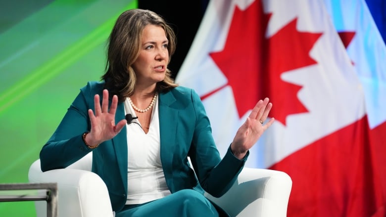 A woman in business attire with a microphone affixed gestures as she sits in a white chair. A Canadian flag is visible behind her.