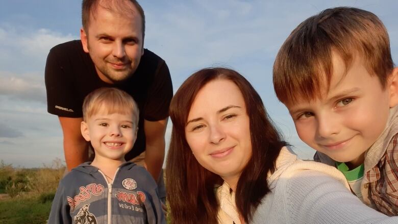 A woman takes a selfie with her two children. Her husband is in the background. 