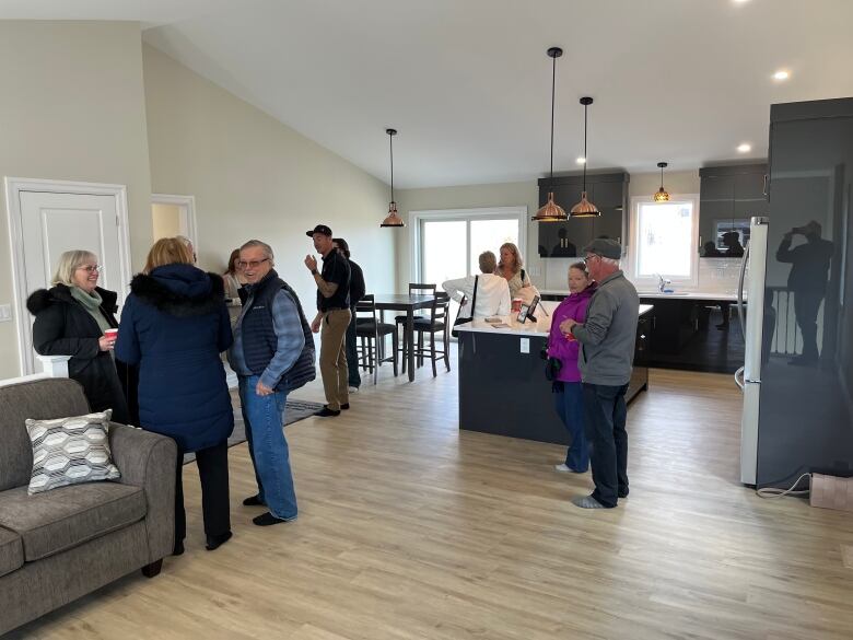 Multiple people stand in a brand new kitchen space. The floors are wooden, and the kitchen cabinets are white. 