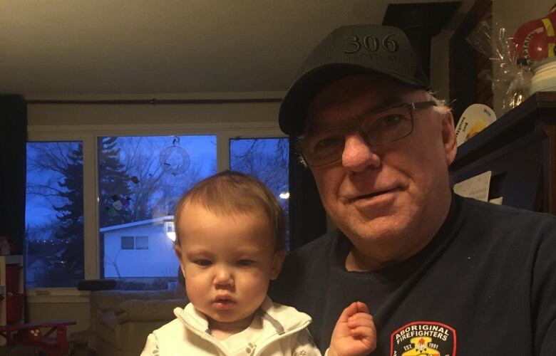 A senior man is wearing a black ball cap that says, '306,' and glasses. A wisp of white hair is seen by his left ear. He is wearing a dark shirt with a firefighting logo on the left breast. He is holding a toddler in his right arm.