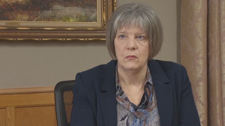 A woman with short grey hair wearing a navy coloured blazer sits on a chair in a boardroom.