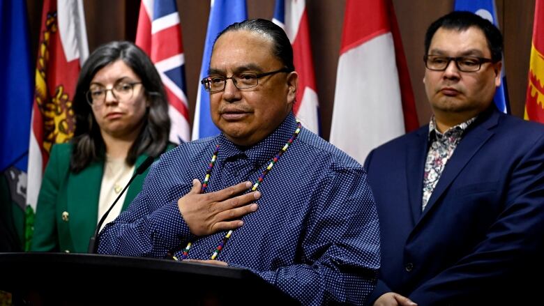 A man in a blue button-up shirt and a beaded medallion around his neck speaks at a podium. Behind him people stand on either side, in front of a wall covered with flags.