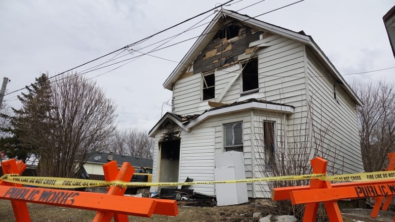 A white house with melted siding and scorch marks near the door is shown behind orange barriers with the words 