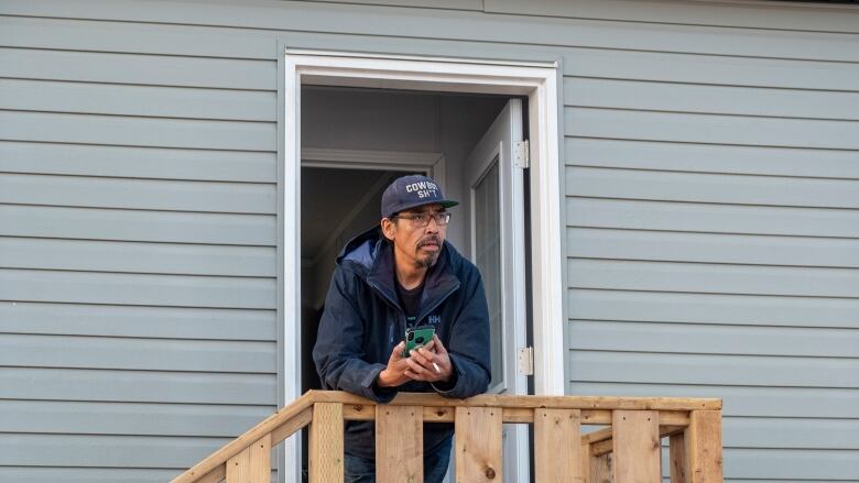 A man stands outside the front door of his house. He's leaning on a railing, looking off into the distance. He's holding his phone and has a cigarette between his fingers. 