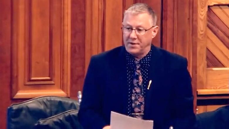 A man with short, grey hair and glasses, wearing a black blazer, black and white polka dot shirt and floral tie, and holding several pieces of paper, speaks.