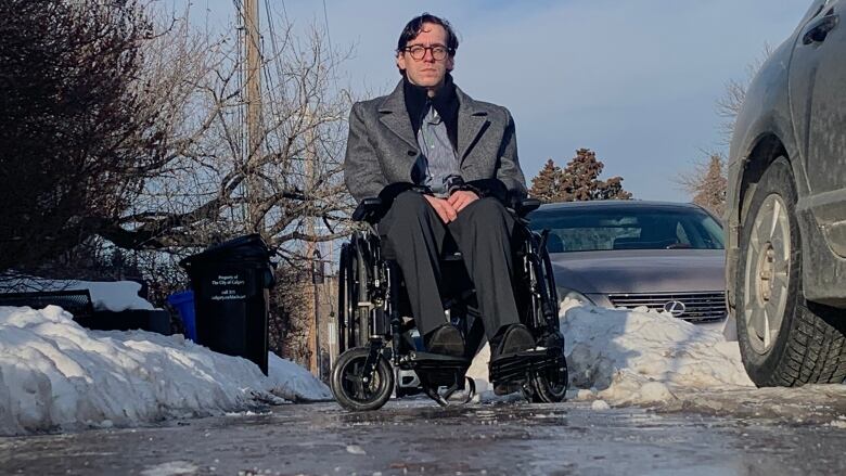 A man in a wheelchair looks at camera while he's on an ice-covered sidewalk, bracketed by snow banks.