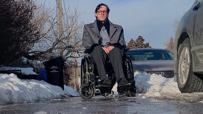 A man in a wheelchair looks at camera while he's on an ice-covered sidewalk, bracketed by snow banks.