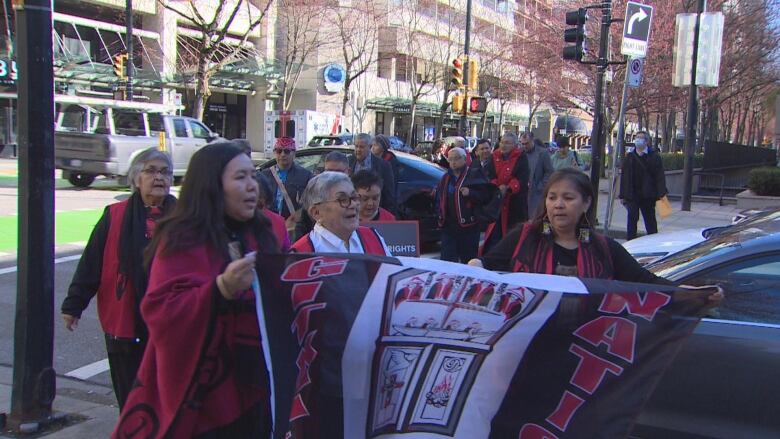 A procession of people holding up a black-and-crimson flag crosses a sidewalk.