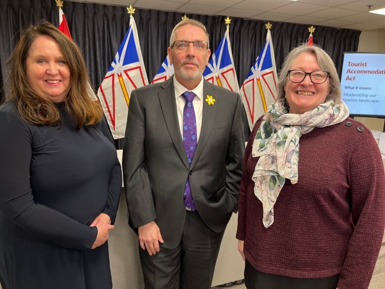 Three smiling people stand in front of a long grey desk. Three Newfoundland and Labrador Flags and two Canada flags are standing on flag poles in the background.