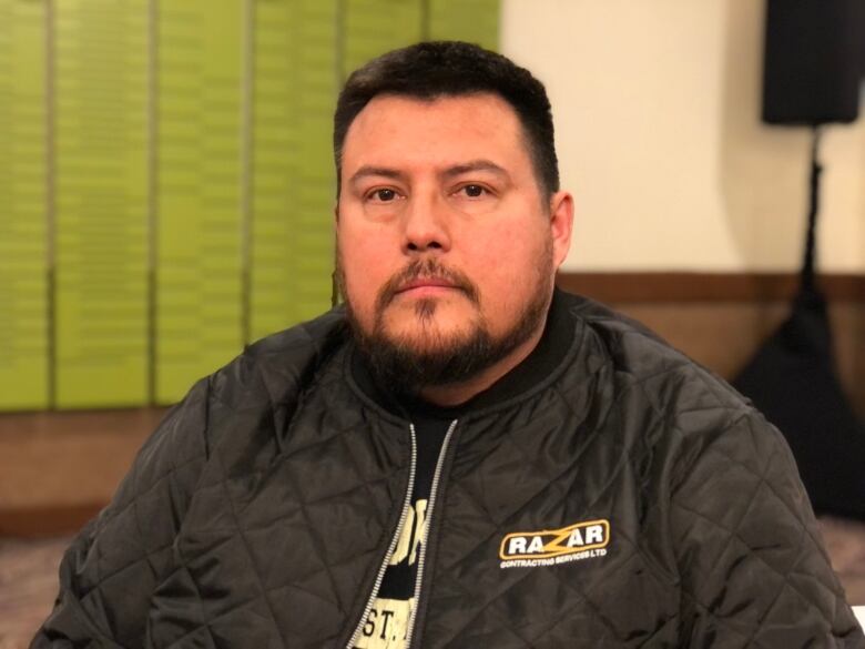 A man with short black hair and short beard in a black jacket looks at the camera.