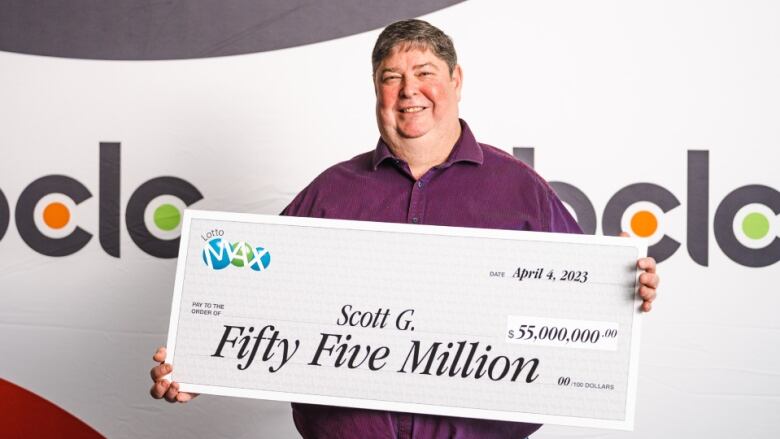 A man stands with a big smile holding an oversized novelty cheque for $55 million.