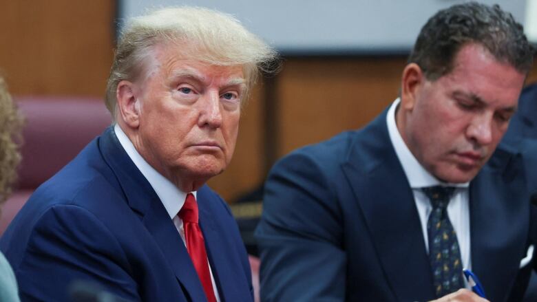 Yellow haired man in blue suit and red tie sits at a courtroom table beside his lawyer looking serious.