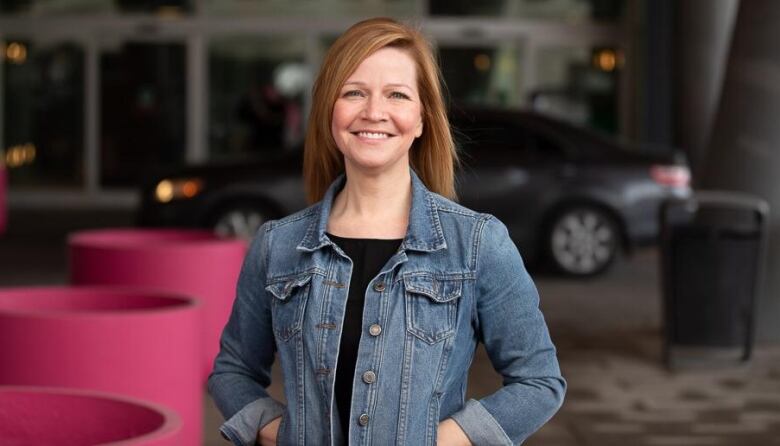 A woman wearing a denim jacket smiles as she poses for a photo with her hands in her pockets. 