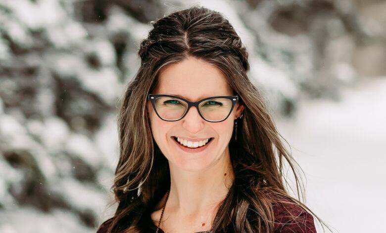 A woman with long brown hair, wearing glasses, smiles as poses for a picture outside in the snow.