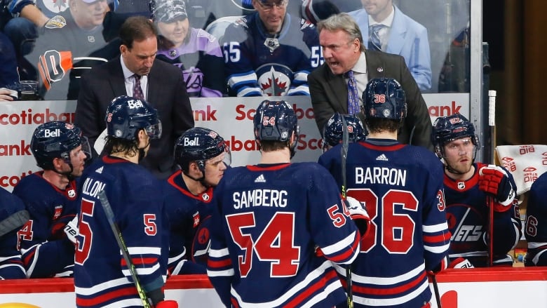 A man with white hair in a grey suit yells at hockey players in blue jerseys as fans watch from behind plexiglass.