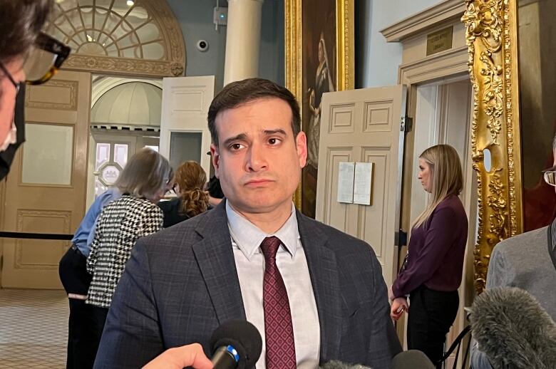 A man with short brown hair wears a grey jacket with a burgundy tie. He is standing in a hallway.