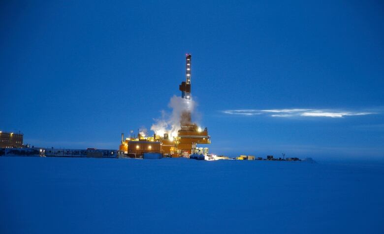 An oil drilling rig is seen at night from a distance on a snowy expanse.