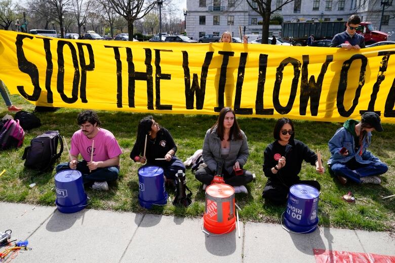 A group of people sit along a sidewalk playing overturned buckets as drums, with a large banner held up behind them, reading, 'STOP THE WILLOW OIL.'