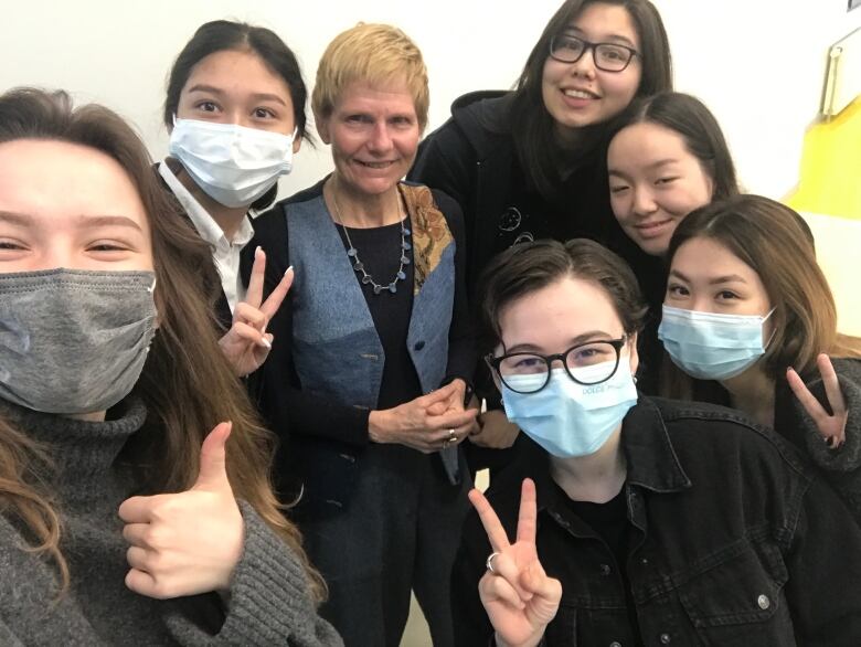 Canadian English teacher pictured surrounded by six female Gr. 12 English students in Almaty, Kazakstan, four of whom are masked and giving thumbs up or peace signs.