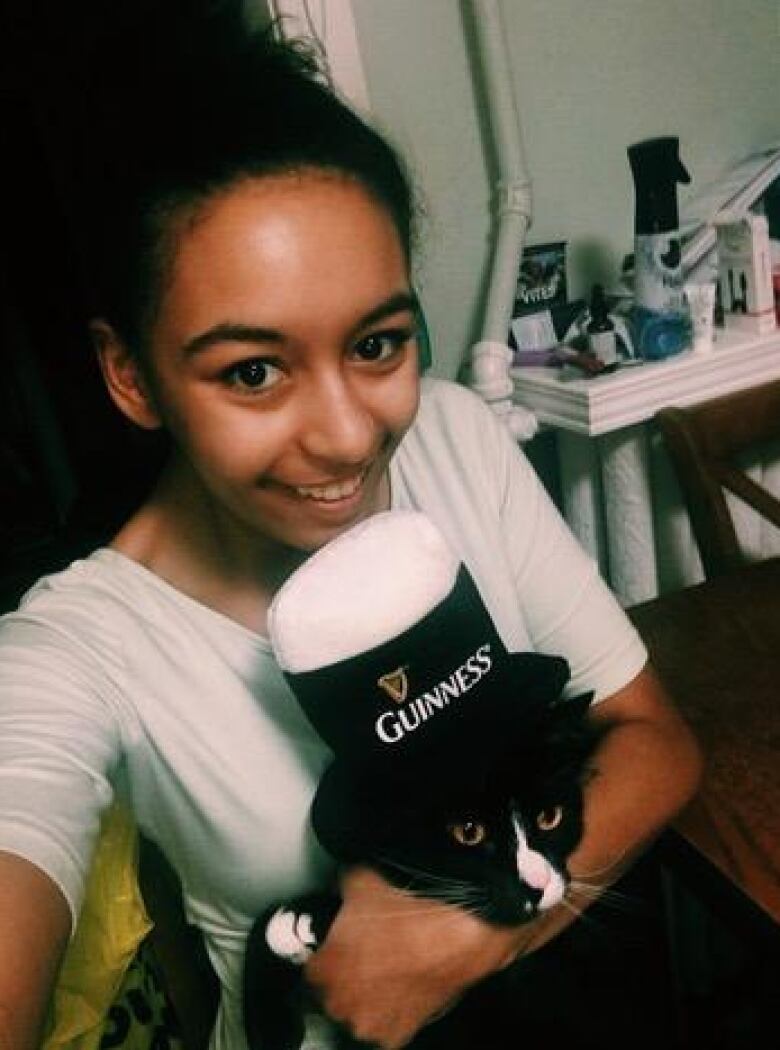 A woman takes a selfie while holding a cat that has a novelty Guinness beer hat on its head. 