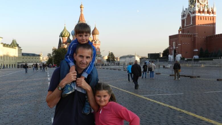 Jason Bronius is shown standing in Moscow's Red Square with his young son on his shoulders and his daughter posing beside him with her hand in her hip. 