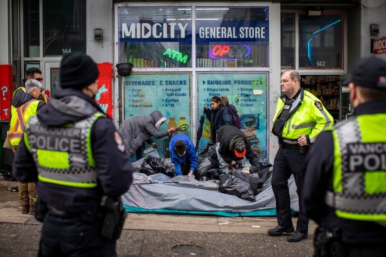 People pack up a tent on the street in front of a convenience store while three police officers watch.