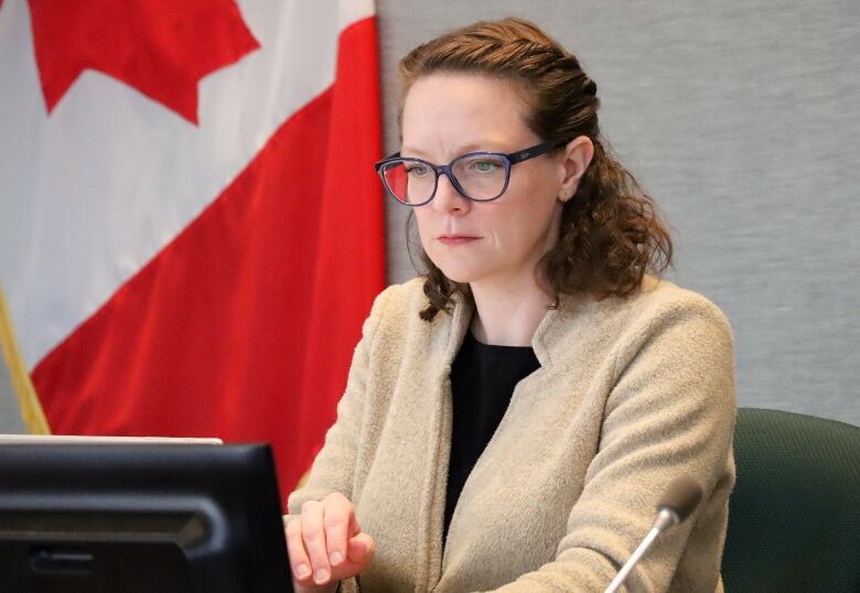 A woman with glasses looks at a computer screen with a Canadian flag in the background.