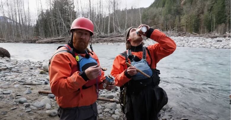 Two men in river rafting gear stand by the edge of the Chilliwack River in B.C. holding cans of beans.