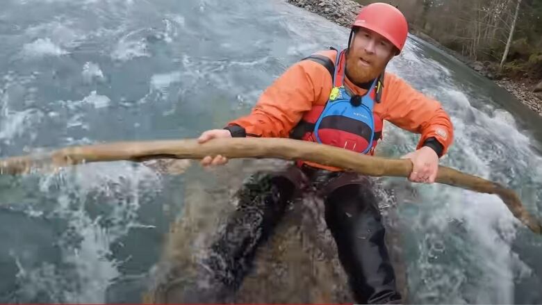 An up-close picture of a red-headed man in river rafting gear holding a branch and sitting on a half-submerged wooden raft in a river.