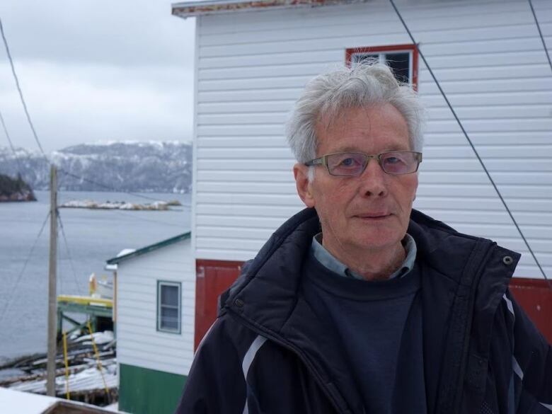 A white-haired man in glasses and a coat stands outside, in front of a red and white building, the ocean in the distance.